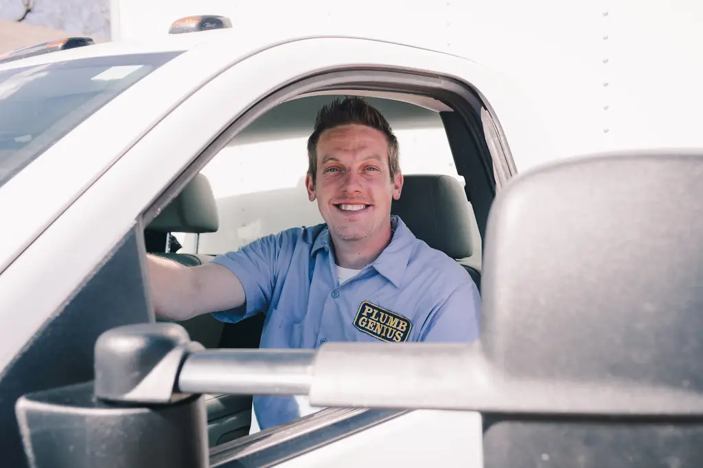 A technician in a Plumb Genius uniform shirt at the wheel of a branded service truck.